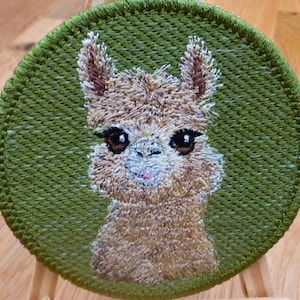 May include: Round embroidered patch with a llama design. The llama has brown fur and large eyes, set against a green background. The patch is suitable for sewing onto clothing or bags. The diameter is approximately 7.6 cm.