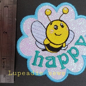 May include: A light blue and pink embroidered patch featuring a cartoon bee and the word "happy" in green. The bee is yellow with black stripes and has a smiling face.