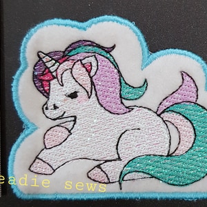 May include: A white unicorn with a pink and teal mane and tail, embroidered on a white cloud-shaped patch with a light blue border. The patch is approximately 5 cm in diameter.