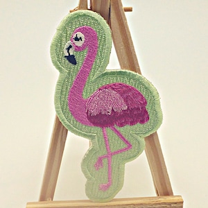 May include: Pink flamingo embroidered patch with a green background. The flamingo is standing on one leg with its head turned to the side.