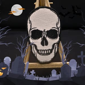 May include: White embroidered skull patch with black eyes and teeth, displayed on a wooden easel in front of a dark background with a graveyard scene.