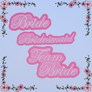 Peut inclure: Trois écussons roses brodés avec du texte blanc en écriture cursive : « Bride », « Bridesmaid » et « Team Bride ». Les écussons ont une bordure décorative et sont posés sur un fond bleu clair, avec des motifs floraux dans les coins.