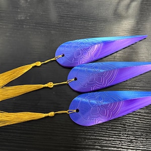 Puede incluir: Tres marcapáginas azules y morados con un diseño floral y borlas doradas. Los marcapáginas tienen forma de lágrima y están hechos de un material liso. Las borlas están unidas con un hilo dorado.