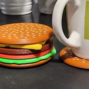 Puede incluir: Una hamburguesa de juguete de capas marrones y blancas con una rebanada de queso amarillo, un tomate rojo y lechuga verde. La hamburguesa está sobre una superficie negra.