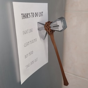 May include: A white piece of paper with the text "Thor's To Do List" written in black. The list includes "Fight Loki", "Clean Mjolnir", "Buy Mead", and "Take Jane Out". A silver and brown hammer is attached to the paper with a brown strap.
