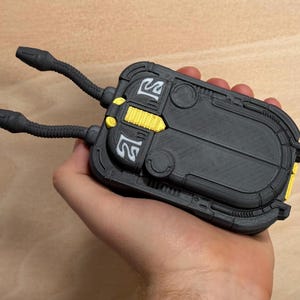 May include: A black and gray device with two flexible, tube-like appendages extending from the top. The device has yellow and white accents, with a textured surface and a rectangular shape. The object is held in a hand.