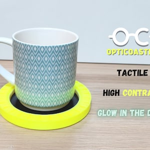 Peut inclure: Un dessous de tasse tactile jaune et noir avec une tasse à motif gris et blanc sur le dessus. Le dessous de tasse est étiqueté "Opticoaster" et porte le texte "Tactile", "Contraste élevé" et "Glow in the Dark".