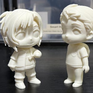 May include: Two off-white, stylized figurines of cartoon characters. One figure is wearing a jacket and holding a small object. The other figure is wearing a t-shirt and has a backpack. The figures are standing on a dark surface.