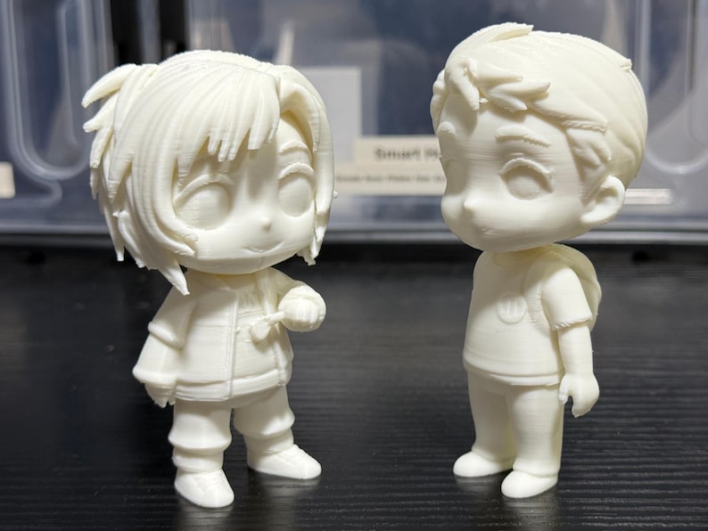 May include: Two off-white, stylized figurines of cartoon characters. One figure is wearing a jacket and holding a small object. The other figure is wearing a t-shirt and has a backpack. The figures are standing on a dark surface.