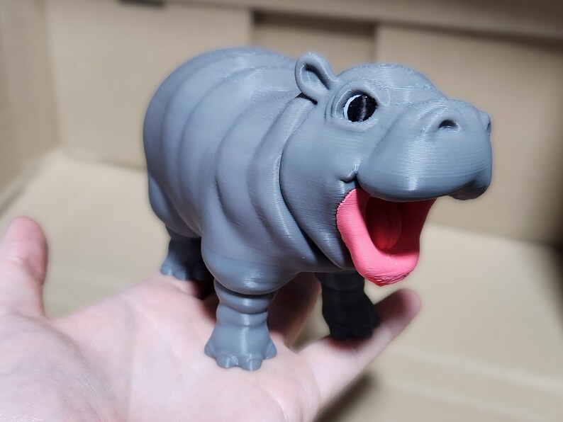 May include: A gray 3D printed figurine of a hippopotamus with an open mouth and a pink tongue. The hippopotamus has a cartoonish appearance with large eyes.