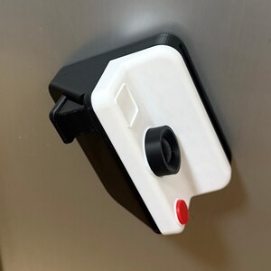 May include: A 3D-printed camera-shaped object with a white front and black casing. It has a black lens, a red button, and a small, square detail on the white surface. The object is mounted on a light grey surface.