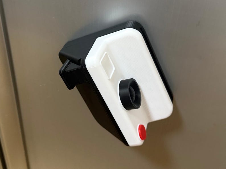 May include: A 3D-printed camera-shaped object with a white front and black casing. It has a black lens, a red button, and a small, square detail on the white surface. The object is mounted on a light grey surface.