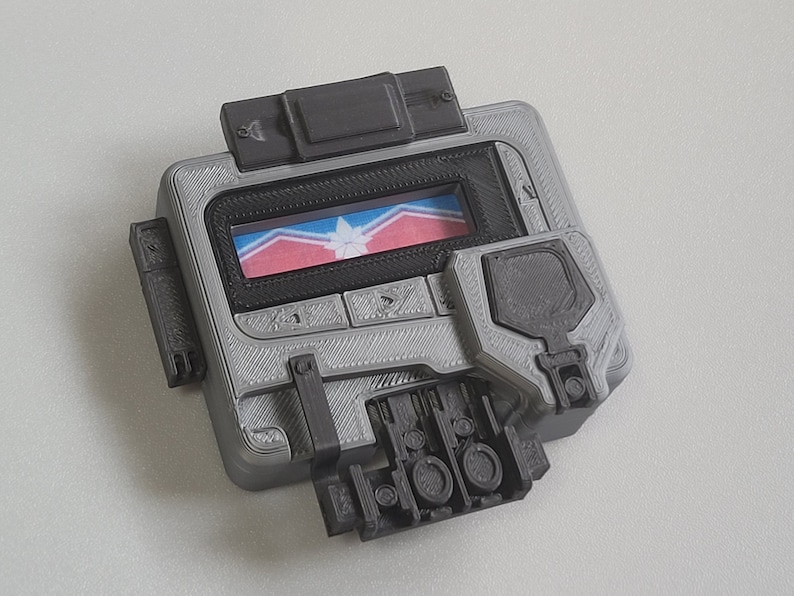 May include: A gray and black handheld device with a small screen displaying a red, white, and blue flag with a star in the center. The device has several buttons and a textured surface.