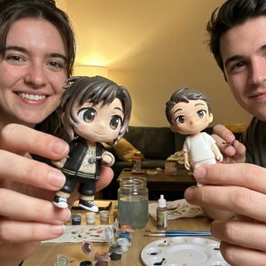 May include: Two anime-style figurines are held up, one in a black jacket and the other in a white shirt. Paint supplies and a palette are on the table. The figurines are being painted.