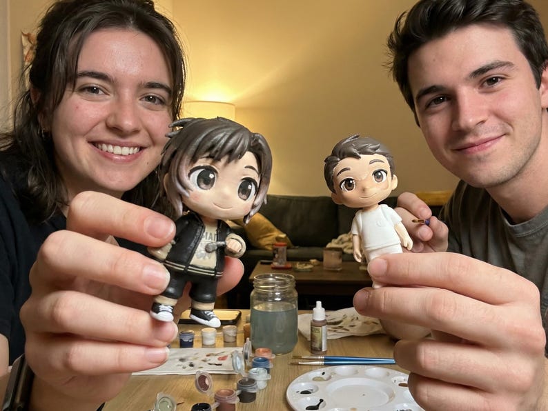 May include: Two anime-style figurines are held up, one in a black jacket and the other in a white shirt. Paint supplies and a palette are on the table. The figurines are being painted.