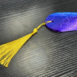 May include: A blue and purple bookmark with a floral design and a gold tassel. The bookmark is made of a solid material and has a hole for the tassel. The tassel is made of gold thread.