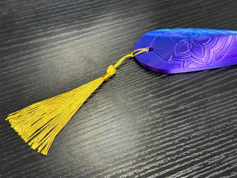 May include: A blue and purple bookmark with a floral design and a gold tassel. The bookmark is made of a solid material and has a hole for the tassel. The tassel is made of gold thread.