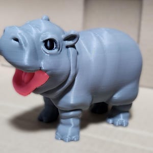 May include: A small, gray, 3D printed hippopotamus figurine with a pink tongue sticking out. The hippopotamus has large, black eyes and a friendly expression.