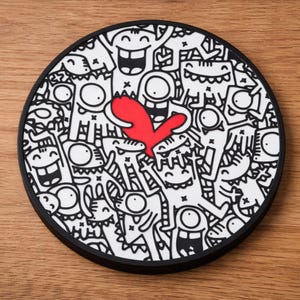 以下が含まれることがあります： 黒い縁取りの丸いコースター。白黒の漫画のキャラクターが描かれた遊び心のあるデザインで、中央に赤いハートがあります。コースターは木製の表面に置かれています。