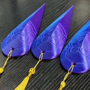 May include: Three teardrop-shaped decorative objects with a gradient of purple to blue. Each has a floral design and a gold tassel. The objects are likely bookmarks or ornaments.