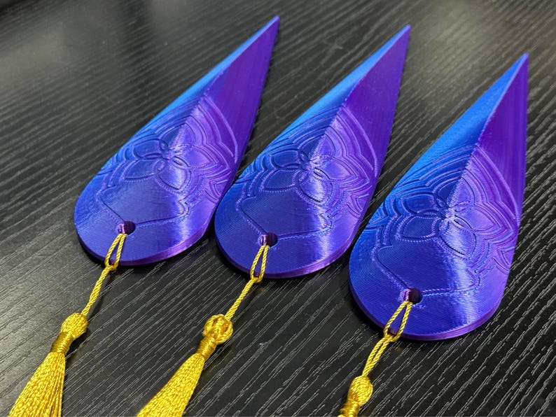 May include: Three teardrop-shaped decorative objects with a gradient of purple to blue. Each has a floral design and a gold tassel. The objects are likely bookmarks or ornaments.