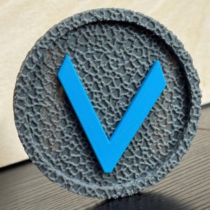 Puede incluir: Un objeto circular gris con una superficie texturizada y un dise&ntilde;o en forma de V azul brillante en el centro. El objeto tiene un borde en relieve y con dibujos. El dise&ntilde;o es simple y moderno.