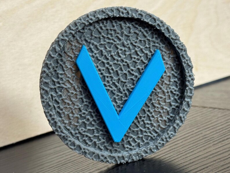 Puede incluir: Un objeto circular gris con una superficie texturizada y un dise&ntilde;o en forma de V azul brillante en el centro. El objeto tiene un borde en relieve y con dibujos. El dise&ntilde;o es simple y moderno.