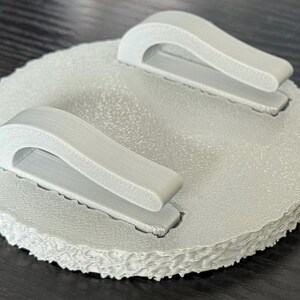 May include: A light gray, circular object with two curved handles. The object has a textured surface and a scalloped edge. The handles are attached to the top surface.