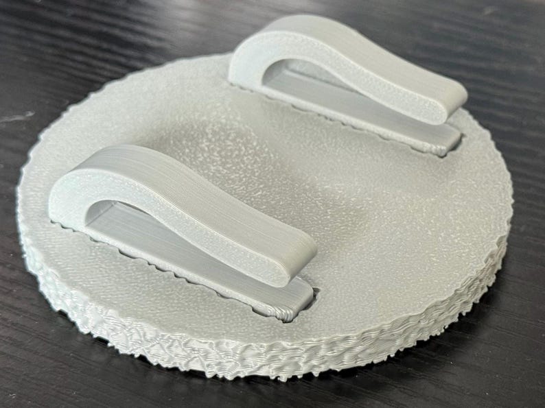 May include: A light gray, circular object with two curved handles. The object has a textured surface and a scalloped edge. The handles are attached to the top surface.