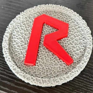 May include: A round, gray coaster with a textured, lace-like pattern. A bright red letter "R" is prominently displayed in the center. The coaster is likely made of a durable material and is designed to protect surfaces from drink rings.