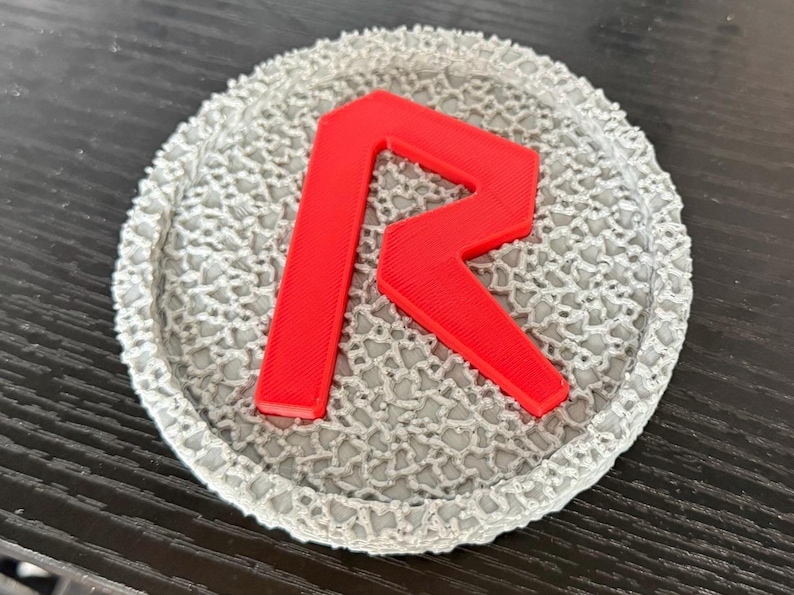 May include: A round, gray coaster with a textured, lace-like pattern. A bright red letter "R" is prominently displayed in the center. The coaster is likely made of a durable material and is designed to protect surfaces from drink rings.
