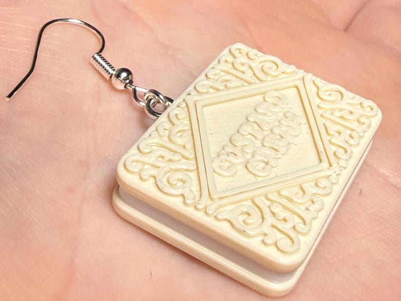 Puede incluir: Pendiente en forma de galleta de color marfil con un gancho plateado. El pendiente presenta un dise&ntilde;o cuadrado con un borde decorativo y un rect&aacute;ngulo central con un dise&ntilde;o de texto apilado. Un accesorio &uacute;nico.