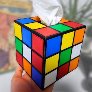 May include: A tissue box holder shaped like a Rubik's Cube with a white tissue peeking out of the top opening. The cube has the classic red, blue, green, yellow, orange, and white colours.