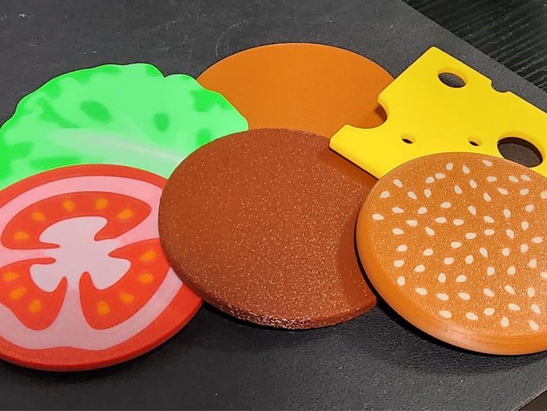 May include: A set of felt food items, including a tomato, lettuce, cheese, hamburger patty, and a sesame seed bun.