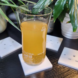 May include: A glass of light yellow juice on a white coaster. The coaster is on a black surface with four white electrical outlet covers.