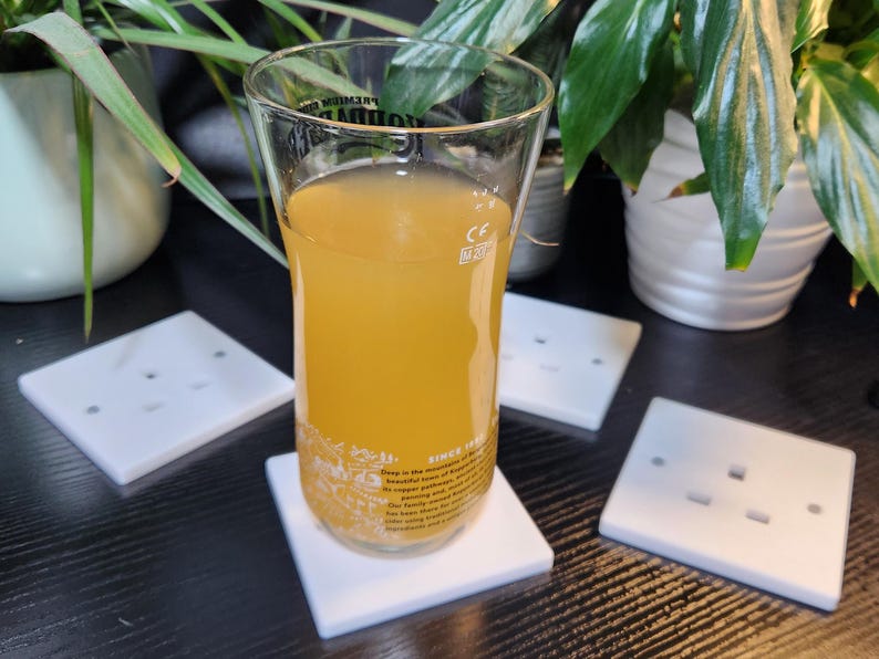 May include: A glass of light yellow juice on a white coaster. The coaster is on a black surface with four white electrical outlet covers.