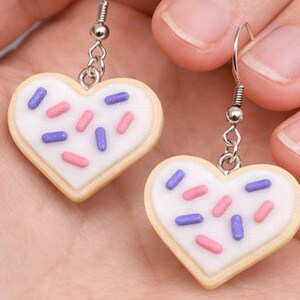 May include: A pair of heart-shaped earrings resembling frosted sugar cookies. Each earring features a white icing base with pink and purple sprinkles, set against a light tan cookie. The earrings hang from silver-colored hooks.