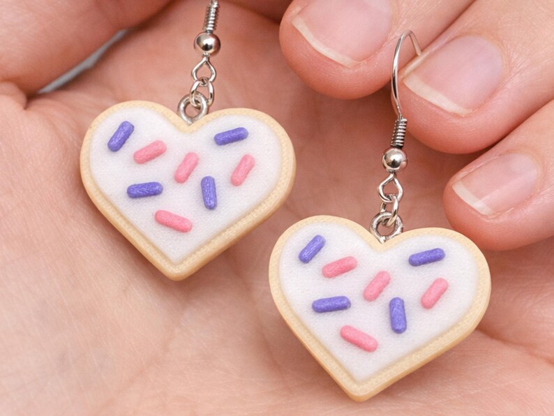 May include: A pair of heart-shaped earrings resembling frosted sugar cookies. Each earring features a white icing base with pink and purple sprinkles, set against a light tan cookie. The earrings hang from silver-colored hooks.
