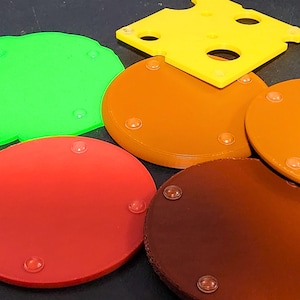 May include: Five round coasters with a textured surface. The coasters are in different colors: green, orange, brown, red, and a darker brown. Each coaster has four small, clear, rubber feet on the bottom.