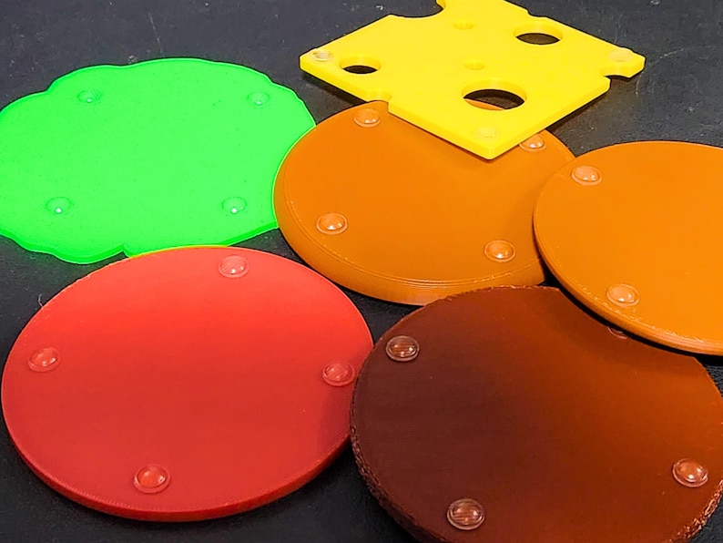 May include: Five round coasters with a textured surface. The coasters are in different colors: green, orange, brown, red, and a darker brown. Each coaster has four small, clear, rubber feet on the bottom.