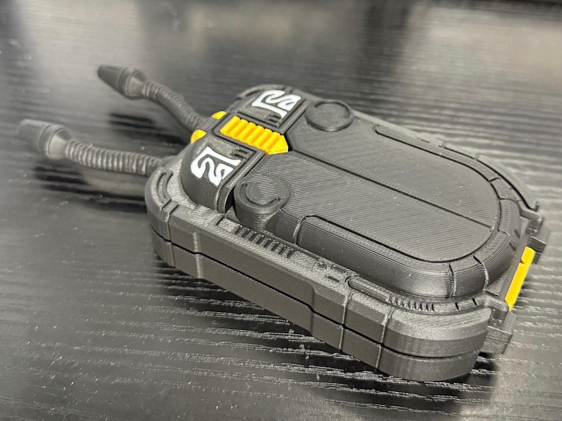 May include: A black and yellow device with two cylindrical tubes extending from the top. The device has a rectangular shape with rounded edges and a textured surface. The design includes yellow accents and white symbols.
