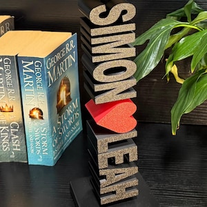 May include: A black wooden bookend with the names "Simon" and "Leah" spelled out in gold glitter letters. A red heart is placed between the names. The bookend is sitting on a black surface in front of a bookshelf with several books.