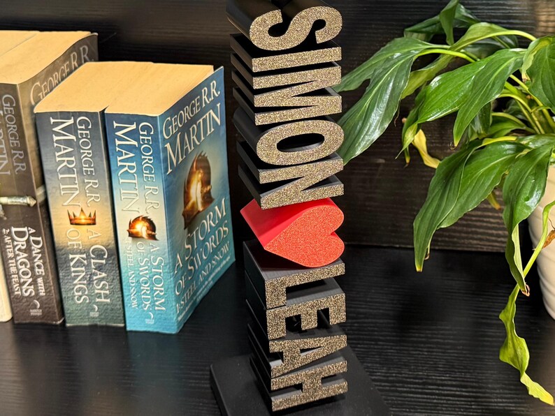 May include: A black wooden bookend with the names "Simon" and "Leah" spelled out in gold glitter letters. A red heart is placed between the names. The bookend is sitting on a black surface in front of a bookshelf with several books.