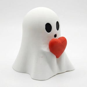 May include: A white ghost figurine with black oval eyes and a surprised expression, holding a bright red heart. The ghost has a draped, sheet-like form, and the heart is positioned in the center.