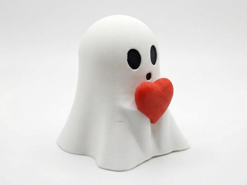 May include: A white ghost figurine with black oval eyes and a surprised expression, holding a bright red heart. The ghost has a draped, sheet-like form, and the heart is positioned in the center.