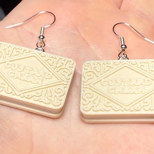Puede incluir: Pendientes de galleta Custard Cream de color crema. Cada pendiente tiene forma rectangular con detalles en relieve y las palabras "Custard Cream". Los pendientes cuelgan de ganchos plateados.