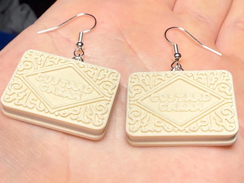 Puede incluir: Pendientes de galleta Custard Cream de color crema. Cada pendiente tiene forma rectangular con detalles en relieve y las palabras "Custard Cream". Los pendientes cuelgan de ganchos plateados.