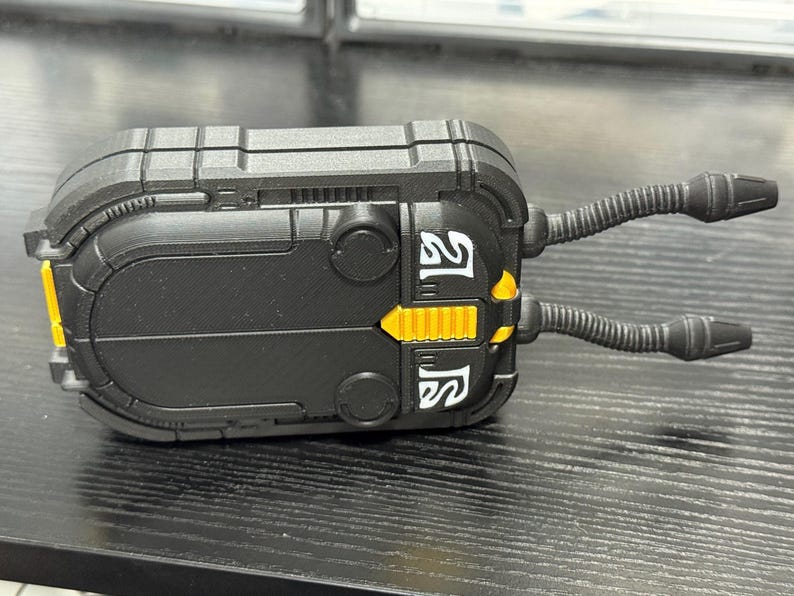 May include: A black, rectangular device with yellow and white accents. Two flexible, tube-like appendages extend from the top. The device has a textured surface and appears to be a piece of equipment or a model.