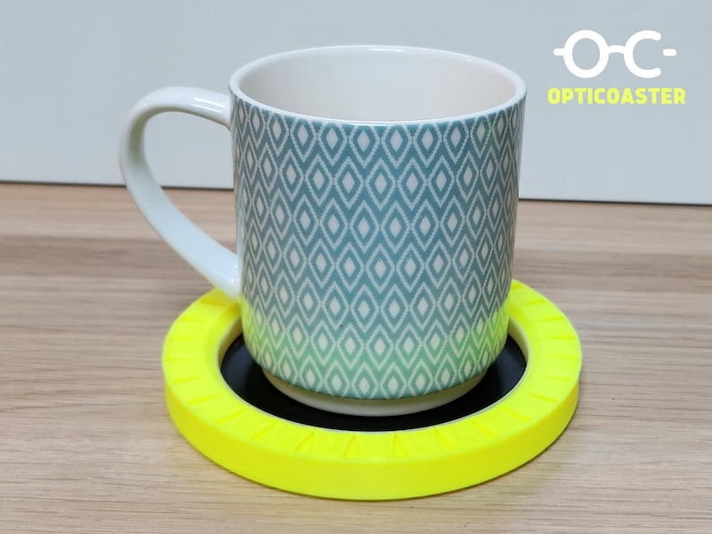 May include: A blue and white patterned ceramic mug sits on a bright yellow circular coaster with a black center. The coaster has a textured, ridged design around the edge. The text "OPTICOASTER" is visible in the upper right corner of the image.