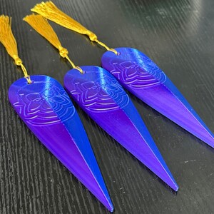 May include: Three gradient blue and purple bookmarks with a floral design and gold tassels. The bookmarks are elongated with a pointed end. The floral design is at the top of the bookmark. The tassels are attached to the top of each bookmark.